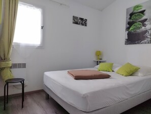Ein Schlafzimmer mit einem Bett, einem Nachttisch mit Lampe, einem kleinen Hocker und einem Fenster mit Vorhängen.