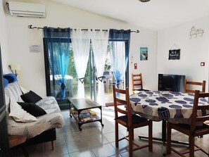 Ein Hotelzimmer mit einer Couch, einem Esstisch mit Stühlen, einem Fernseher und Blick ins Freie durch ein Fenster mit durchsichtigen Vorhängen.