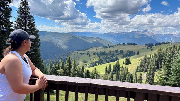 A person standing on a wooden deck overlooking a mountainous landscape with trees and a clear sky.