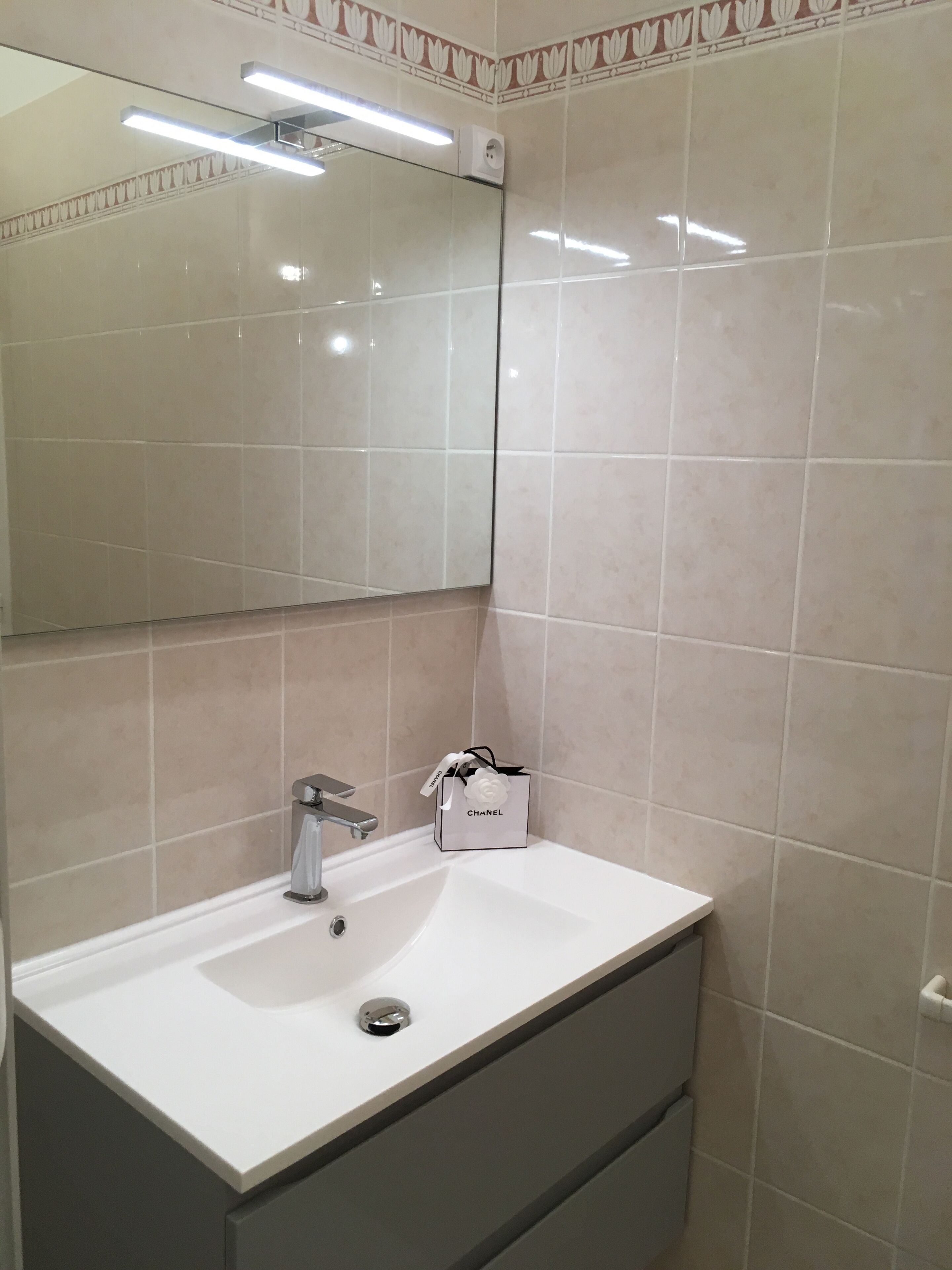 Un baño con un lavabo blanco, un espejo y un bolso Chanel sobre el mostrador.
