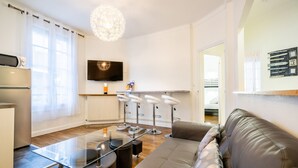 A modern living room with a sofa, a glass coffee table, a TV mounted on the wall, and a kitchen area with a refrigerator and a microwave. - ⭐️⭐️⭐️Cathédrale, bright space in the heart of town (Reims)
