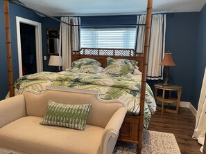 A four-poster bed with a tropical leaf pattern bedspread, a wooden chair with a green pillow, and a small table with a lamp.