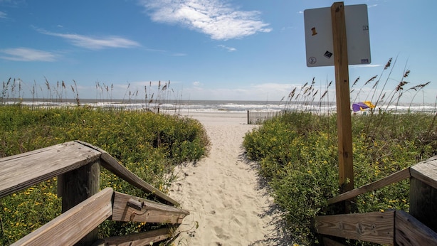 Una pasarela de madera que conduce a una playa, con un cartel indicador en primer plano.