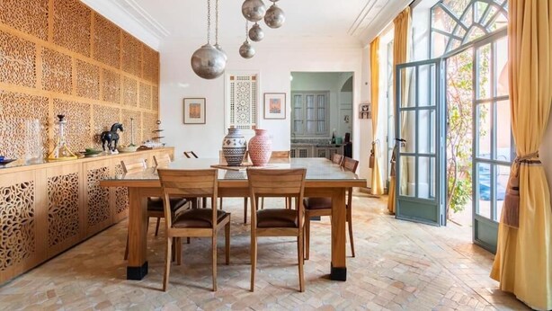 Een eetruimte met een houten tafel en stoelen, een decoratieve wand en een groot raam met gordijnen.