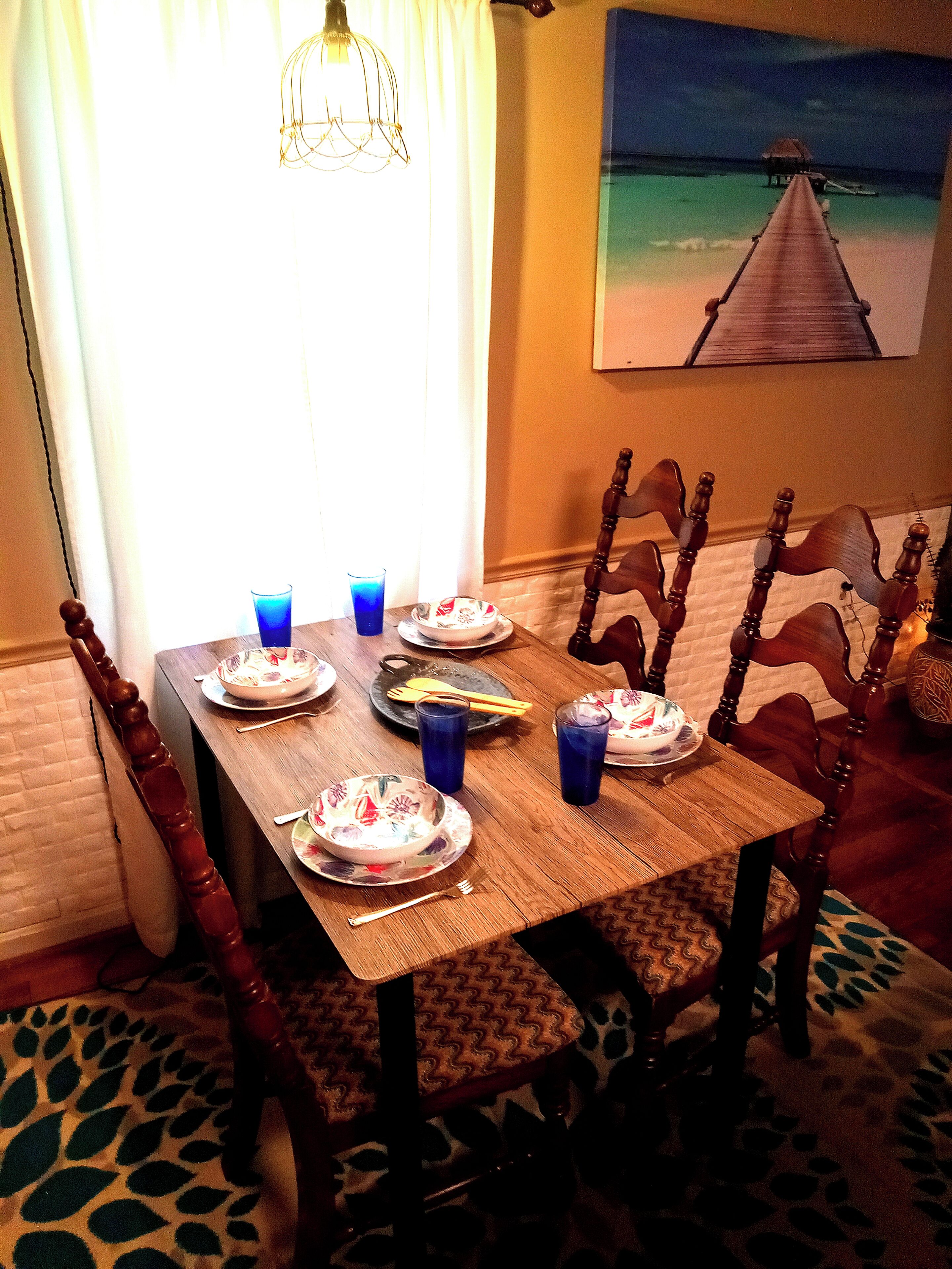 A dining table set with plates, bowls, and blue glasses, surrounded by wooden chairs with patterned cushions.