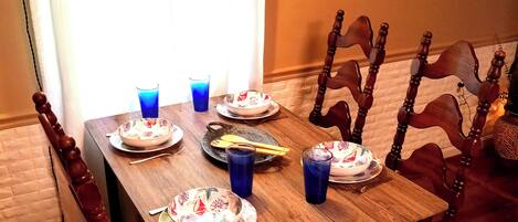 A dining table set with plates, bowls, and blue glasses, surrounded by wooden chairs with patterned cushions.