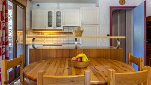 A wooden dining table with chairs, a fruit bowl, and a kitchen area with white cabinets and a clock on the wall. - Village Center, Near Ski Slopes, Courses, and Shops (La Clusaz)