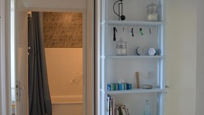 A bathroom with a shower, a toilet, and a sink. There is a shelf with various items including books, a bottle, and a small wooden stool.