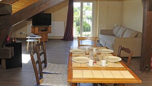 Een eetgedeelte met een houten tafel gedekt voor vier personen, een televisie op de achtergrond en een balkon met uitzicht op groen.