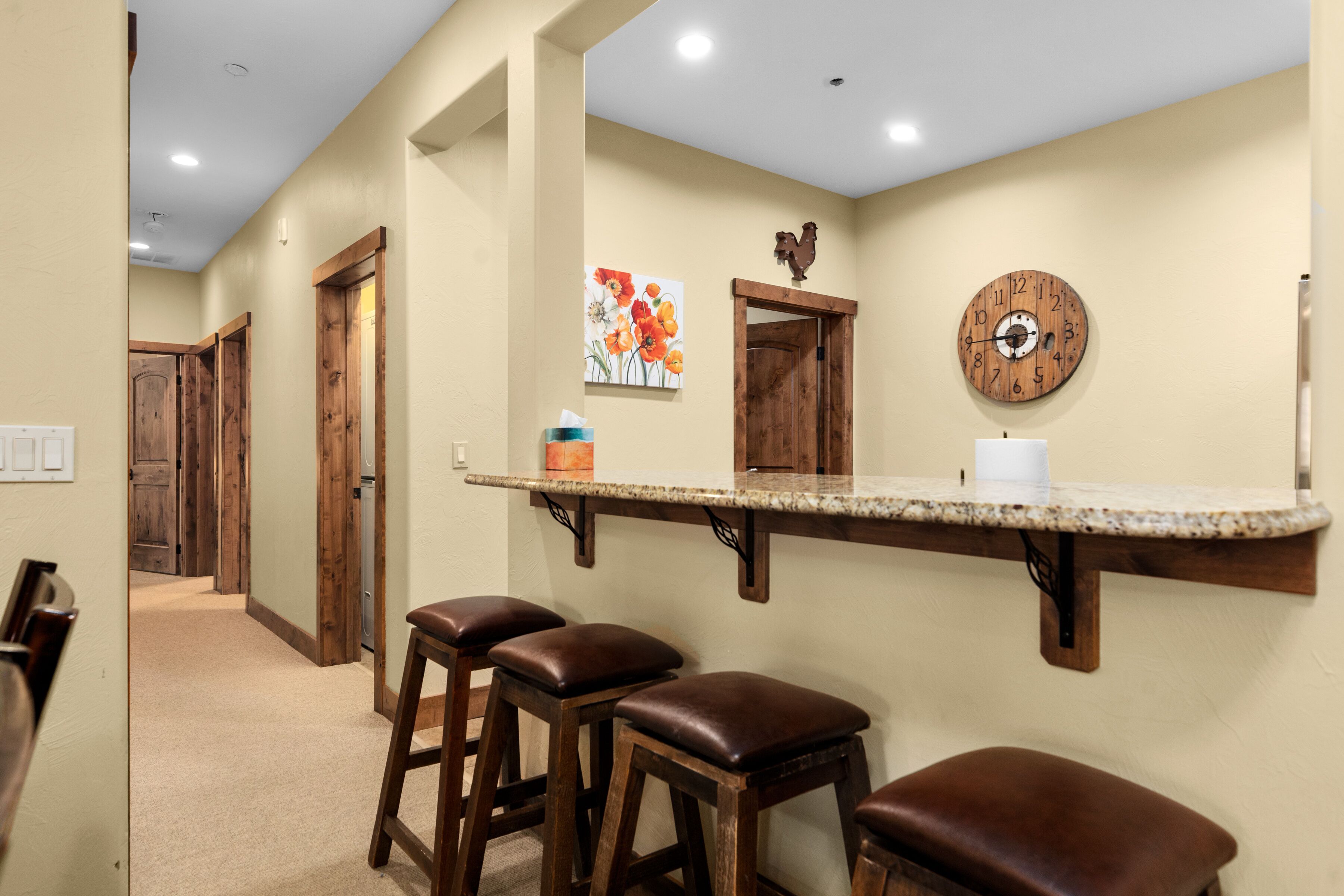 A hallway with wooden doors, a kitchen area with a bar counter, stools, and a clock on the wall.
