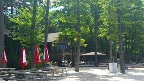 Un parque con mesas de picnic y sombrillas rojas, rodeado de árboles y un edificio al fondo.
