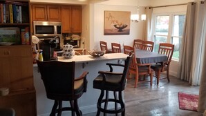 Una cocina con armarios de madera, microondas, cafetera y zona de comedor con sillas y mesa de madera.