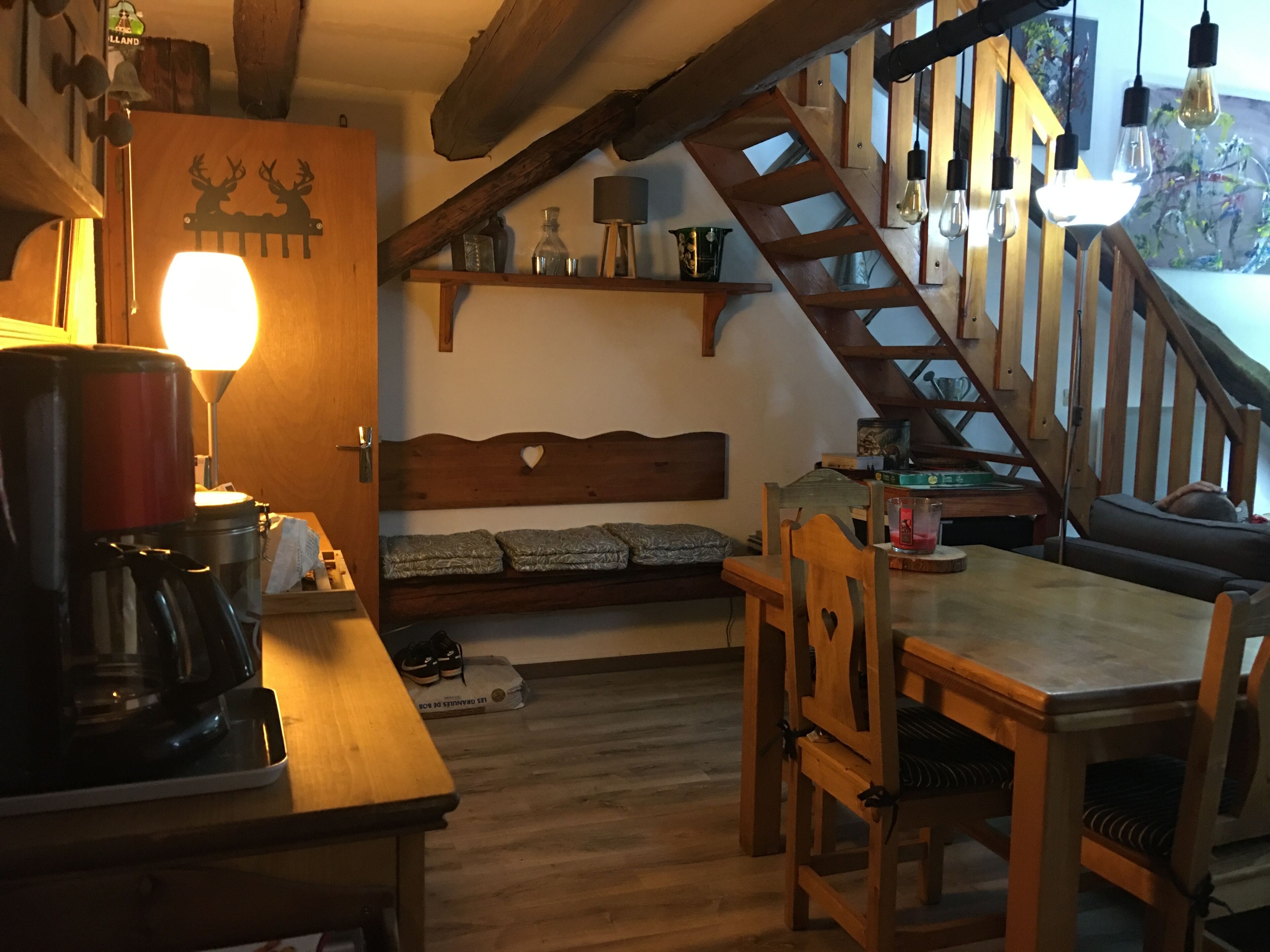 A cozy room with a wooden table, chairs, a bench, a staircase, and a wall with shelves.