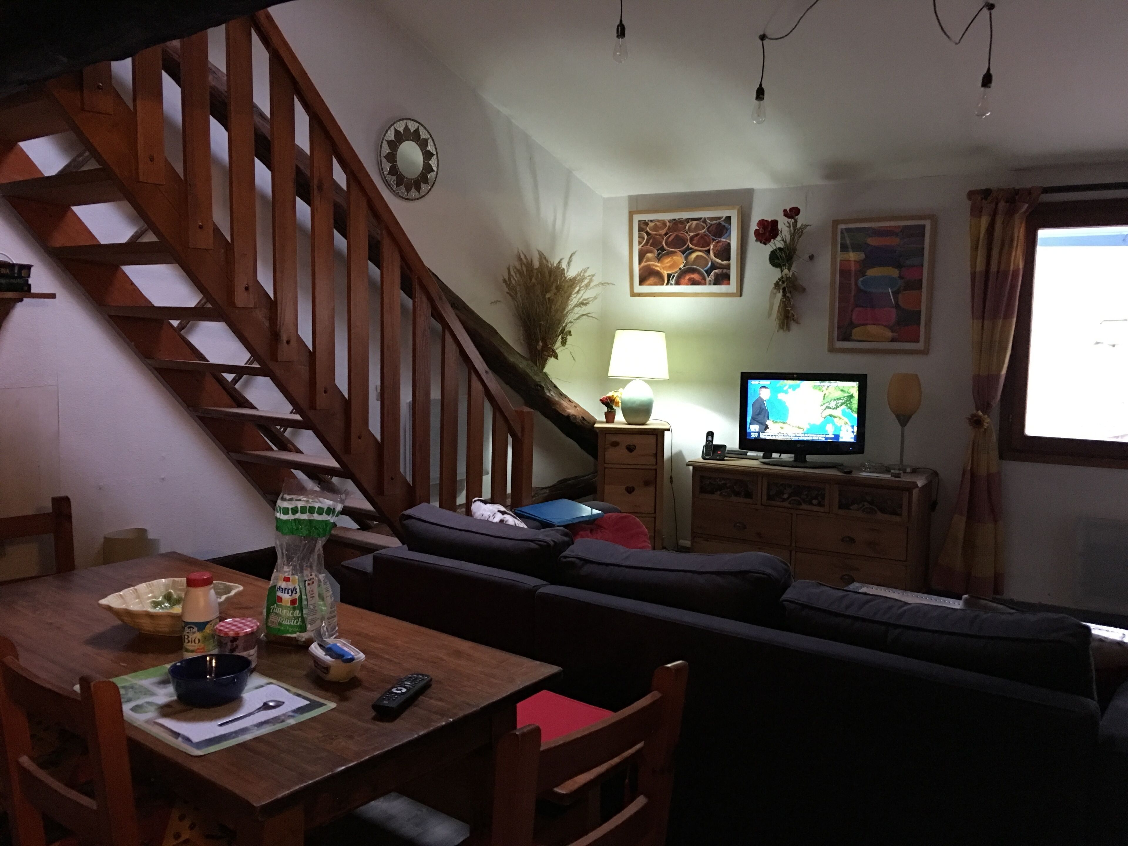 A cozy living room with a wooden staircase, a television, and a dining area with a table set for two.