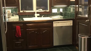 A kitchen with dark wooden cabinets, a stainless steel dishwasher, and a window above the sink. - JACARANDA.  A comfortable, secure two-story colonial.  Whole house.  (Milton)