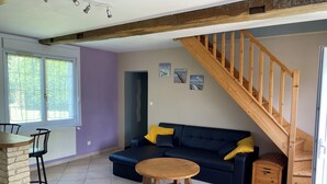 Un salón acogedor con una escalera de madera, un sofá azul con cojines amarillos, una mesa de comedor de madera y una zona de cocina con ventana.