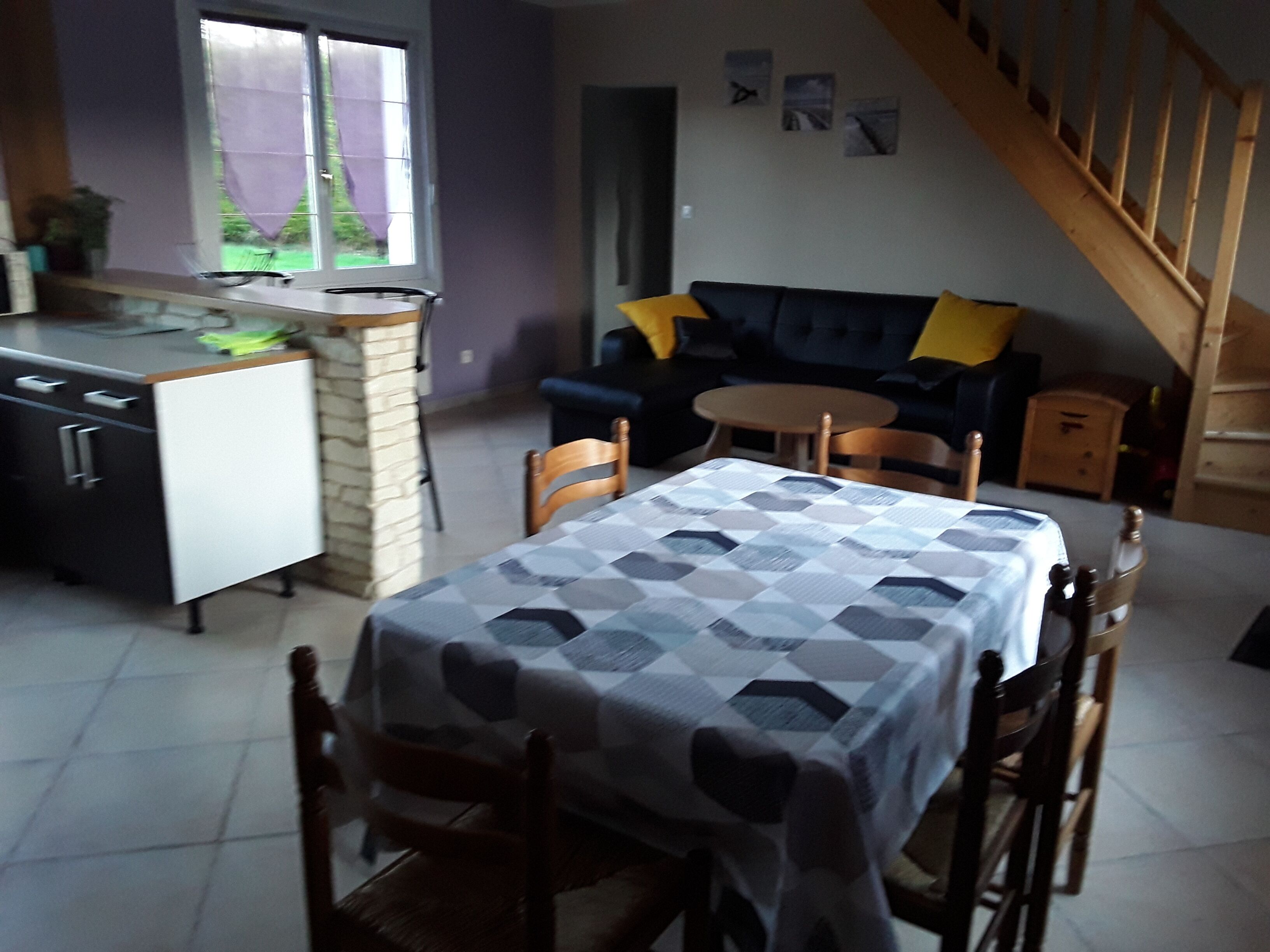 A dining area with a table covered by a patterned tablecloth, surrounded by wooden chairs. A kitchen area with a stove and cabinets is visible to the left.