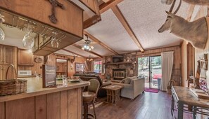 Interior rústico de cabana com paredes de madeira, lareira de pedra, área de refeições e bar com adega suspensa.