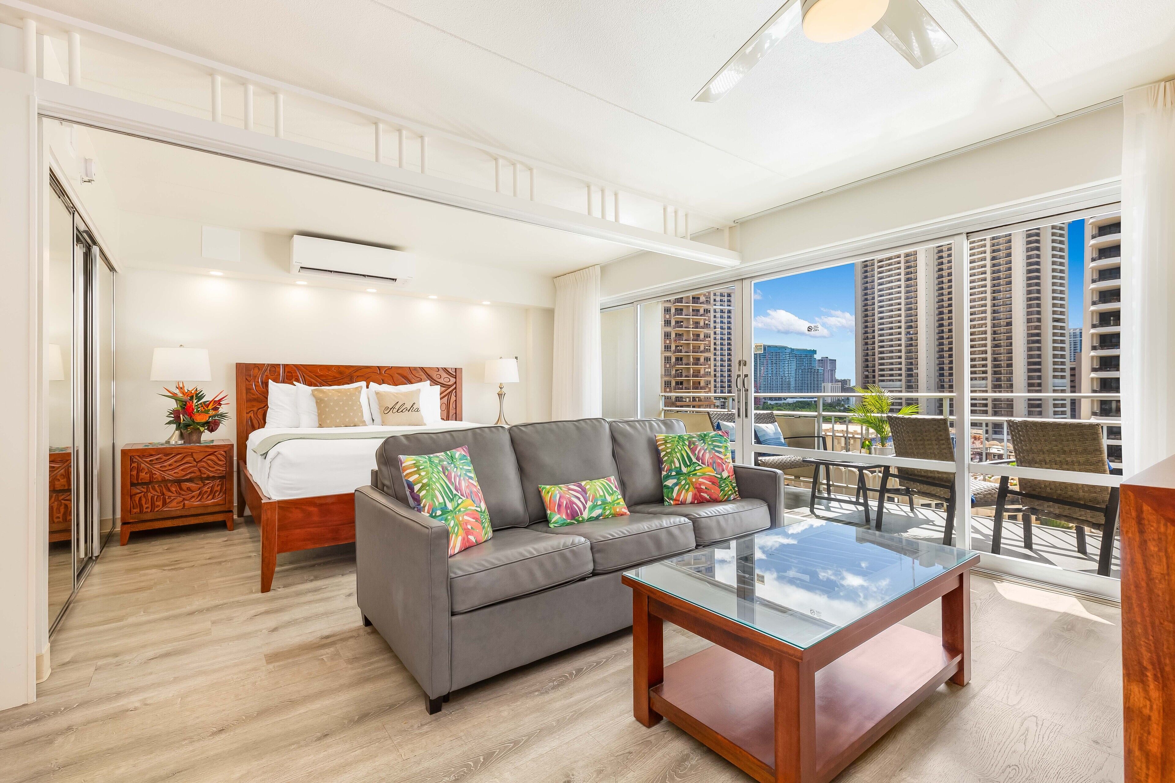 A hotel room with a bed, a gray sofa, a glass coffee table, and a balcony with a city view.
