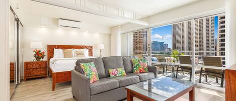 A hotel room with a bed, a gray sofa, a glass coffee table, and a balcony with a city view.
