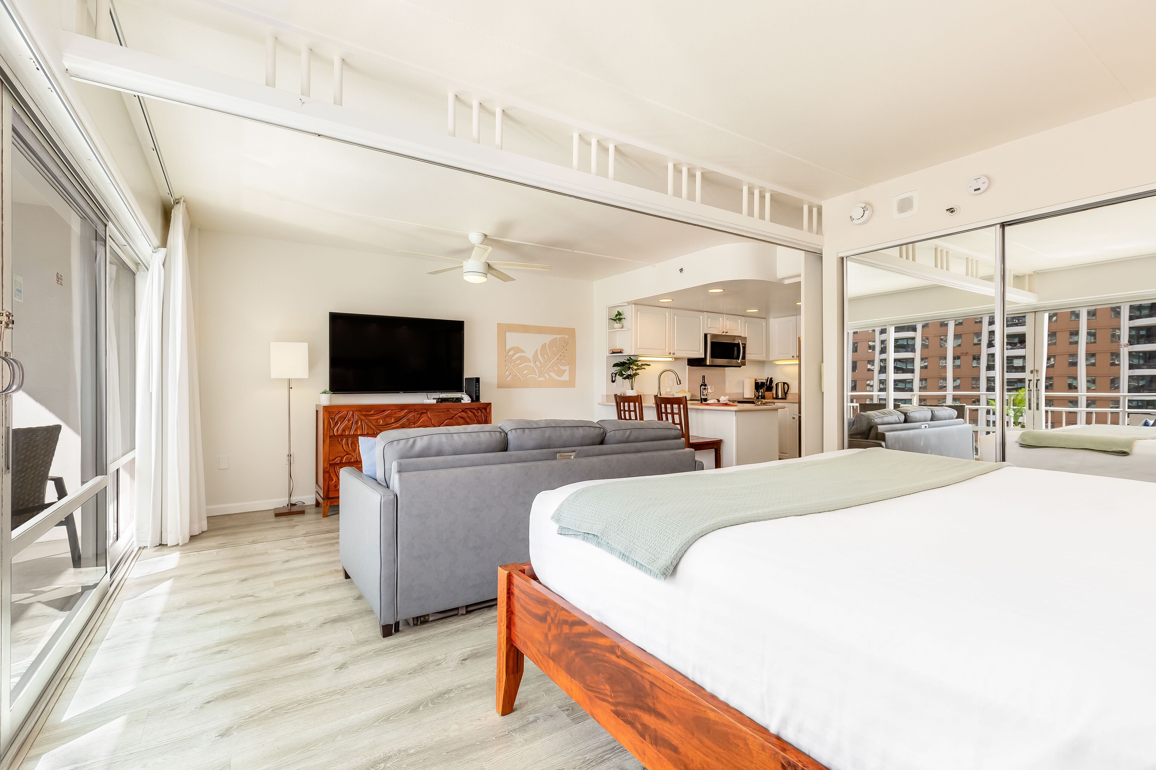 Habitación de hotel con cama, sofá, televisor, ventilador de techo y vista del paisaje urbano a través de amplias ventanas.