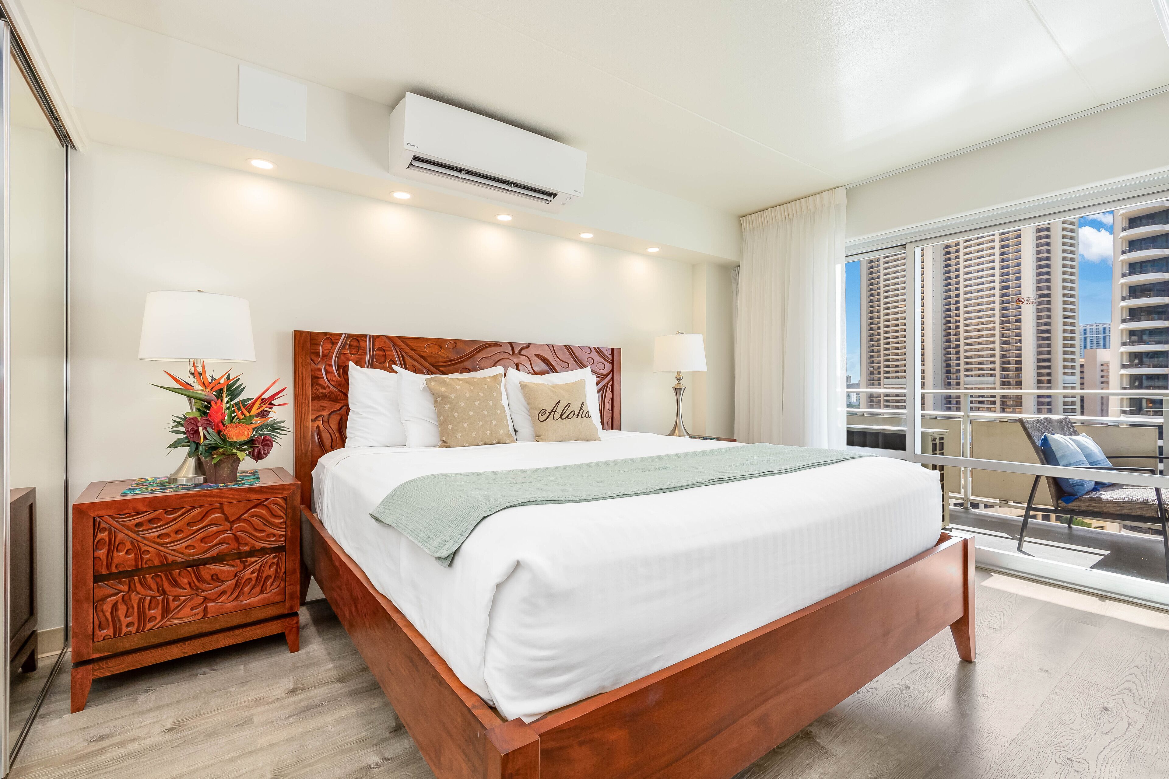 Une chambre d’hôtel avec un grand lit, une table de chevet, une plante en pot, une fenêtre donnant sur la ville et un appareil de climatisation.