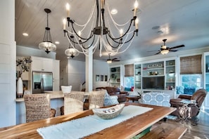 A spacious living area with a wooden dining table, wicker chairs, a brown leather sofa, a ceiling fan, and a chandelier.