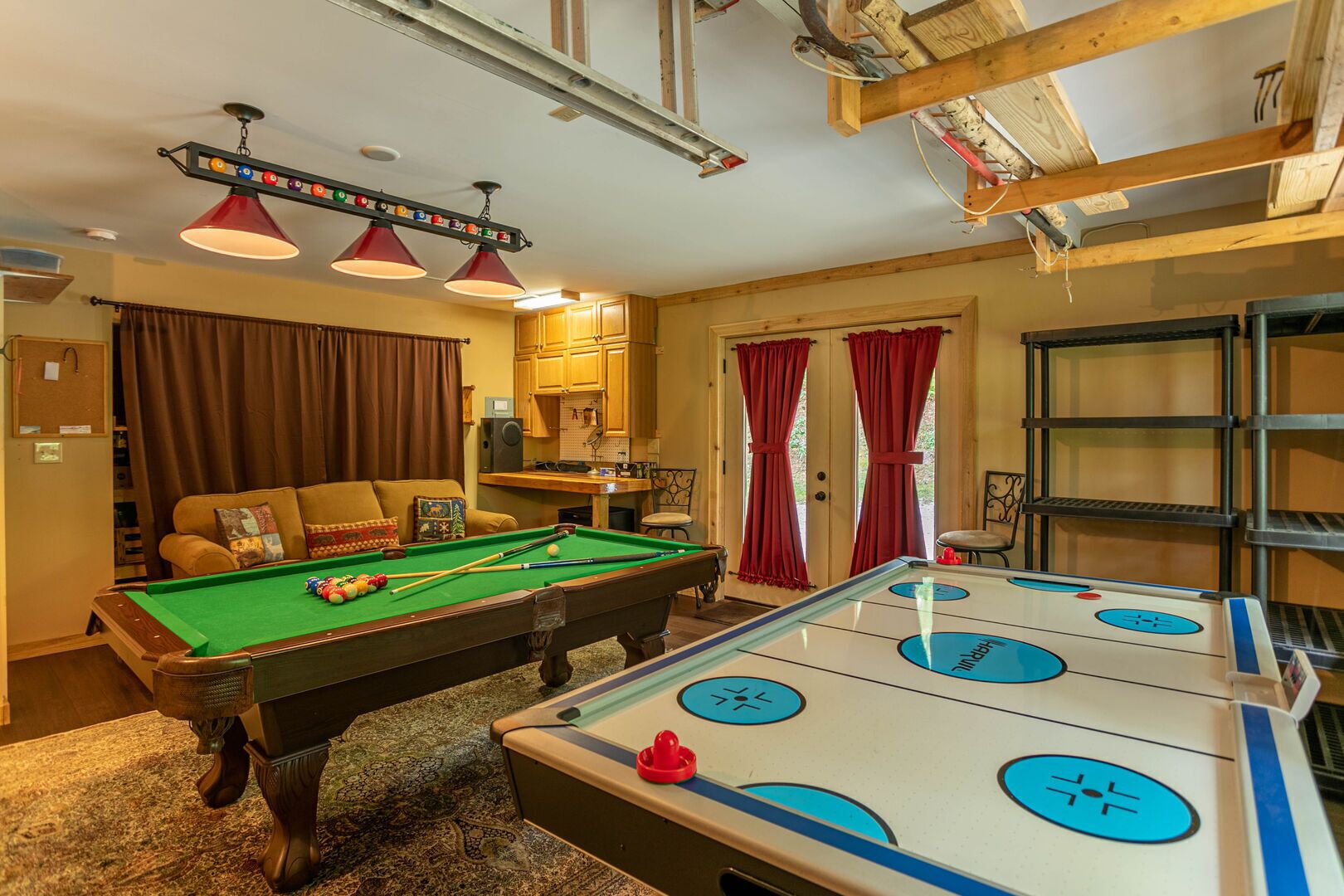 A pool table with a foosball table, a sofa, and a speaker in a room with wooden beams and red curtains.