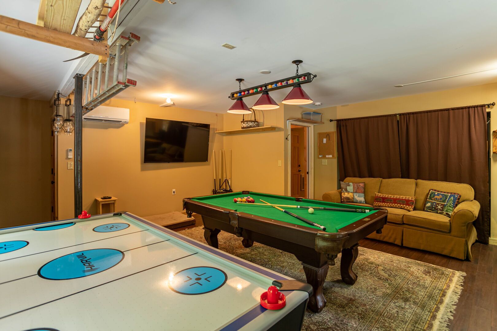 A pool table with cues and balls, a foosball table, a sofa, and a television in a room with wooden floors and a brown curtain.