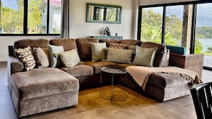A spacious living room with a large sectional sofa, a small round table, and large windows offering a view of the outdoors. - Walk to Sunset Cocktails, Lake Access and Birdwatching - Arenal peninsula (de Nuevo Arenal)