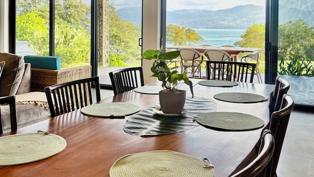 A dining area with a wooden table, chairs, and a potted plant. Large windows offer a view of greenery and a distant landscape. - Walk to Sunset Cocktails, Lake Access and Birdwatching - Arenal peninsula (de Nuevo Arenal)
