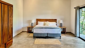 A bedroom with a wooden headboard, a bed with white bedding, two bedside tables with lamps, and a large window with a view of greenery. - Walk to Sunset Cocktails, Lake Access and Birdwatching - Arenal peninsula (de Nuevo Arenal)