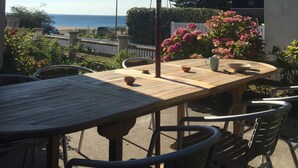 Un patio al aire libre con mesa y sillas, un paraguas grande y vista al océano.