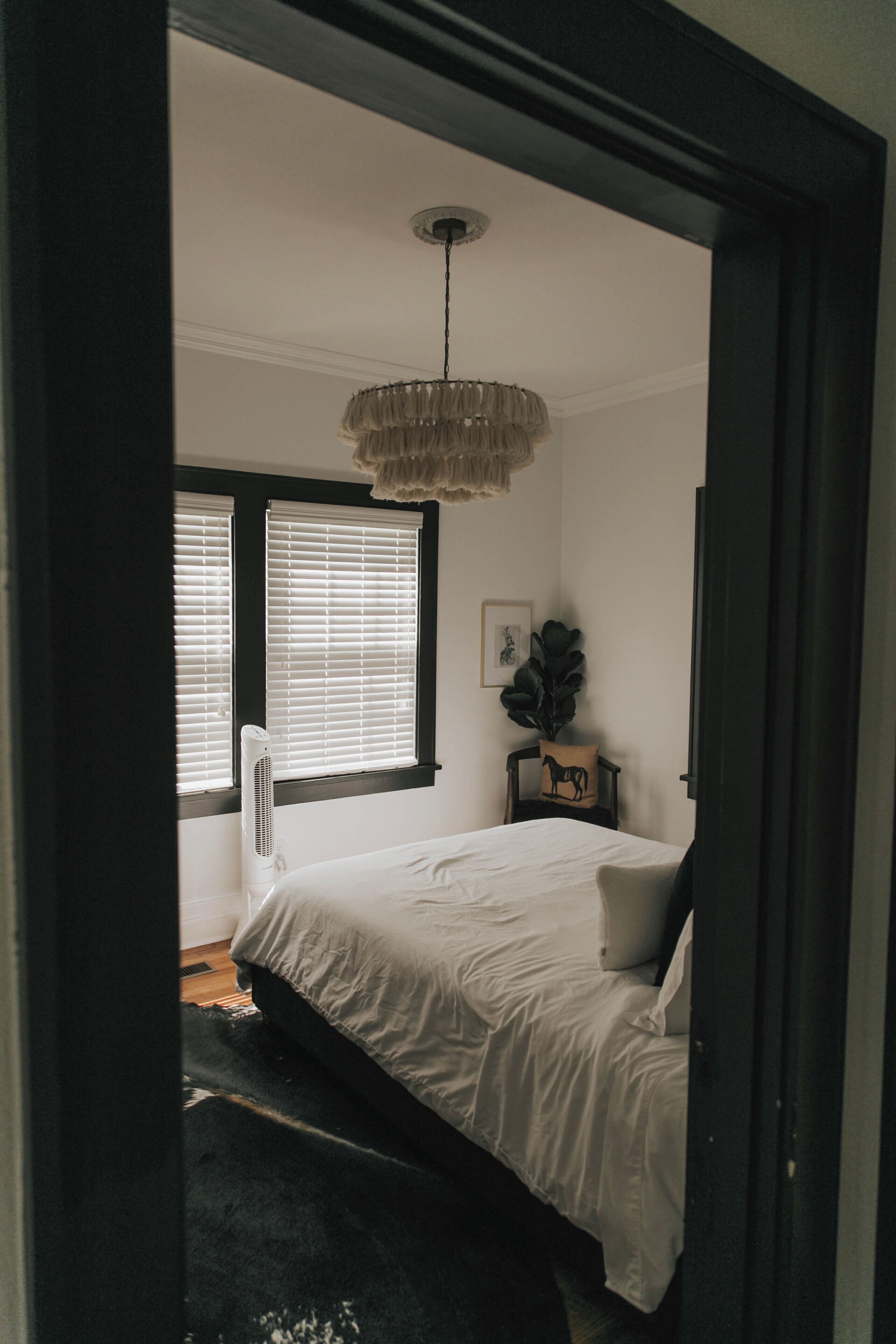 A bedroom with a bed, a fan, a window with blinds, and a plant on a side table.