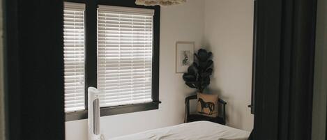 A bedroom with a bed, a fan, a window with blinds, and a plant on a side table.