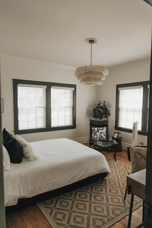 Une chambre avec un grand lit, deux fenêtres équipées de stores, un luminaire suspendu, une chaise avec un coussin et une petite table sur laquelle est posée une plante.