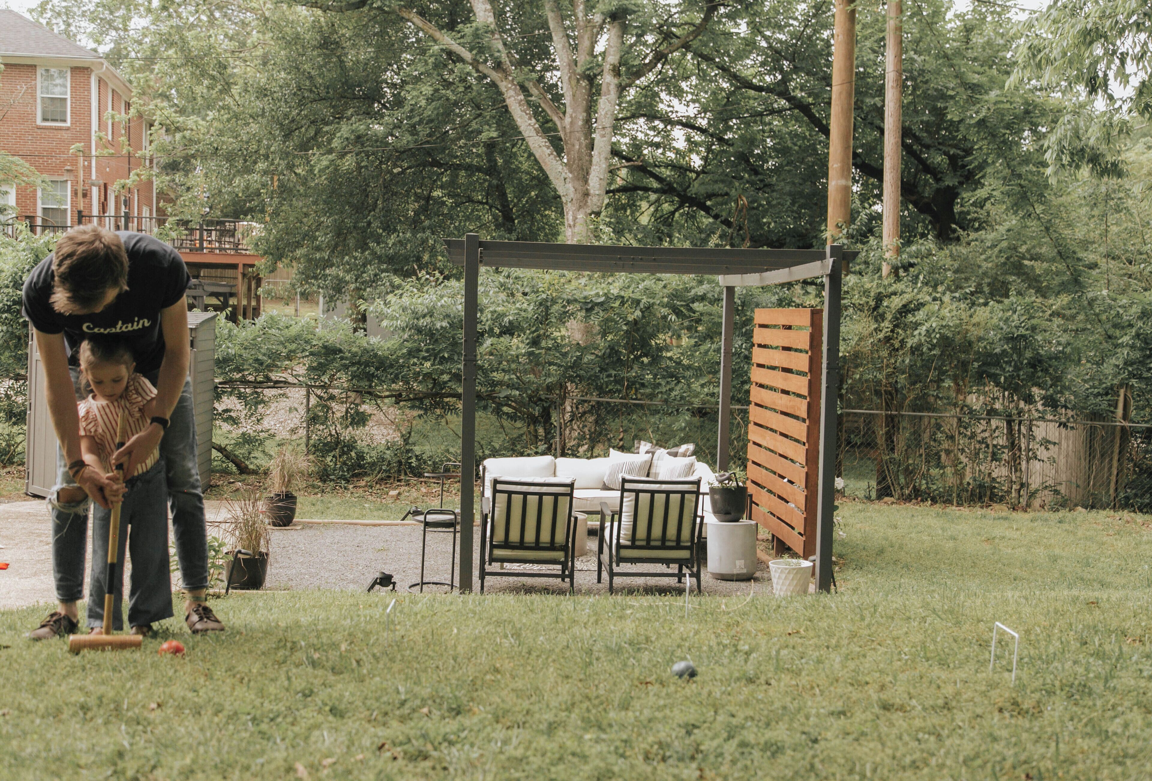 A covered outdoor seating area with a table and chairs, surrounded by a garden and a house in the background.