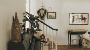 Un salón acogedor con escalera, suelo de madera, una planta, un sillón de mimbre y un cuadro en la pared.