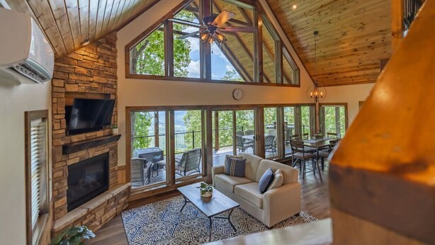 A spacious living area with a stone fireplace, large windows, a ceiling fan, and a dining area with outdoor seating. - Red Rock Vista Cabin-Fall Family Vacation in Jasper (Jasper)