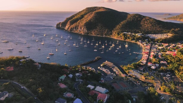 Un pueblo costero con un puerto deportivo lleno de veleros, rodeado de colinas y un pequeño puerto.