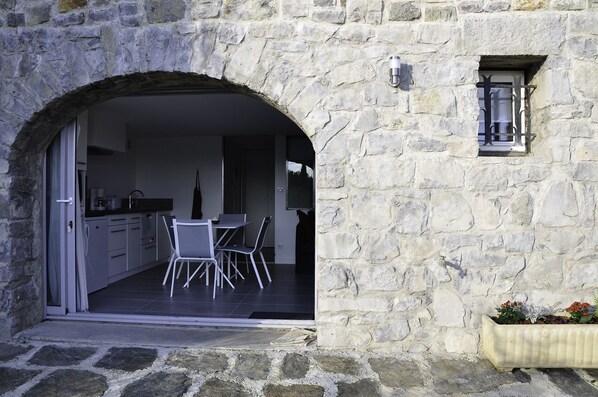 Un portail en pierre en arc donne accès à une cuisine moderne avec des meubles blancs et à un coin repas comprenant une table et des chaises.