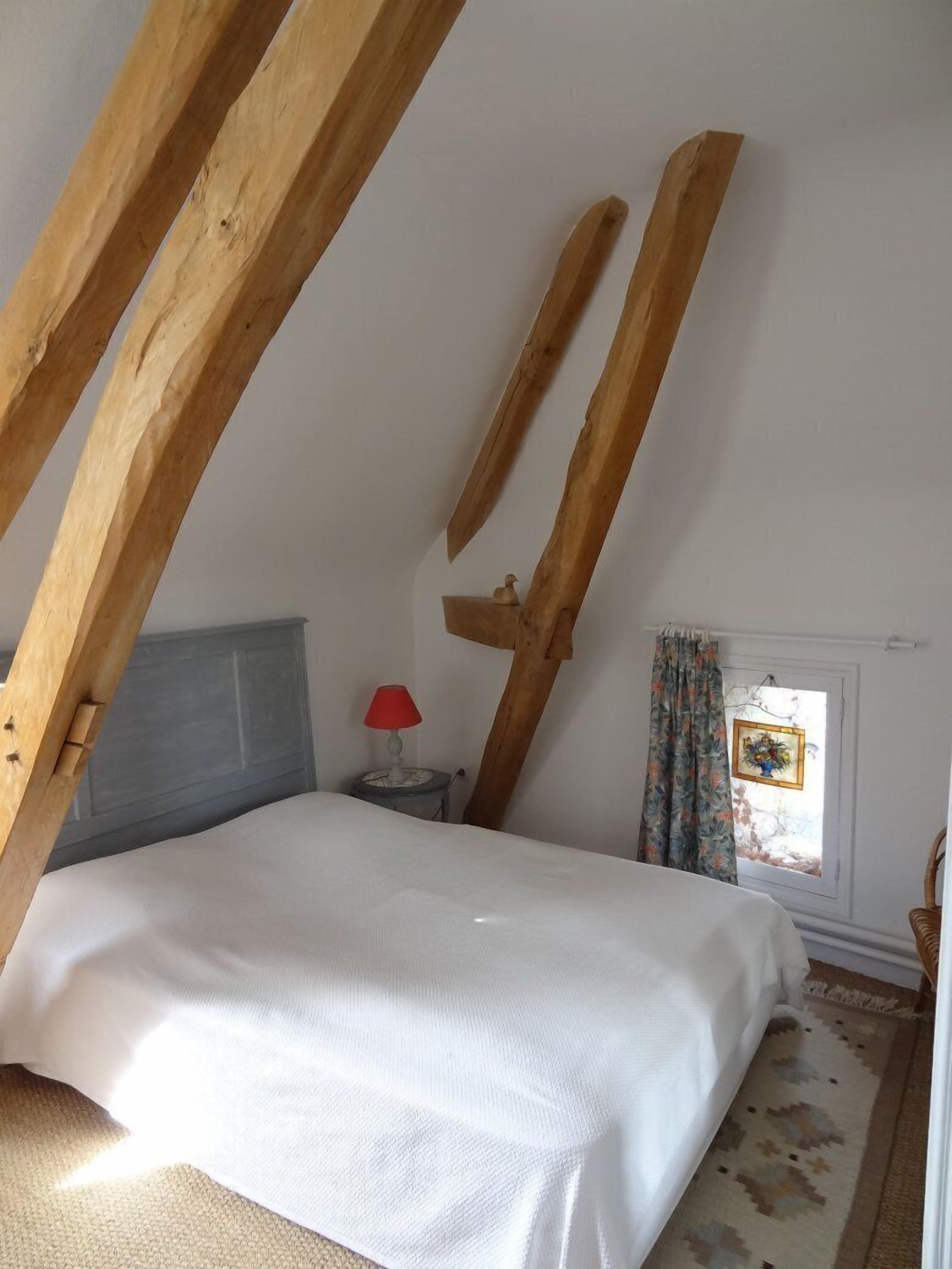 Une chambre avec un plafond en pente, un lit, une table de chevet avec une lampe, une fenêtre avec des rideaux et un petit cadre photo.