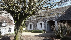 Un edificio con un gran árbol en primer plano, un muro de piedra y un camino pavimentado que lleva a una puerta blanca con contraventanas.