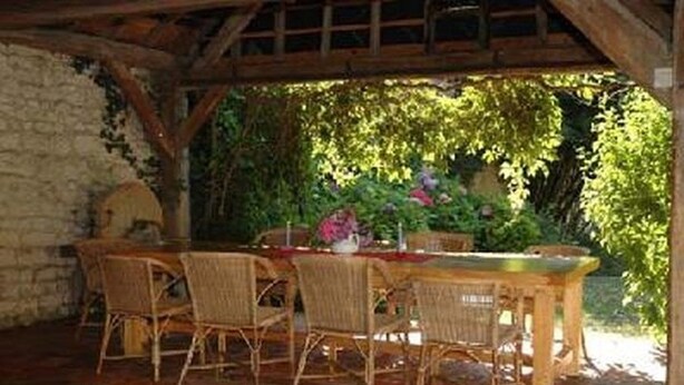 Un área de comedor al aire libre con muro de piedra, pérgola de madera y abundante vegetación.