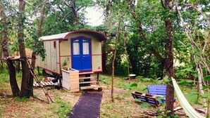 A small cabin with a blue door and a red trim, surrounded by trees and a black path leading to it. - Gypsy Caravan and Wooden Cabin In Nature (Saint-Méard-de-Gurçon)