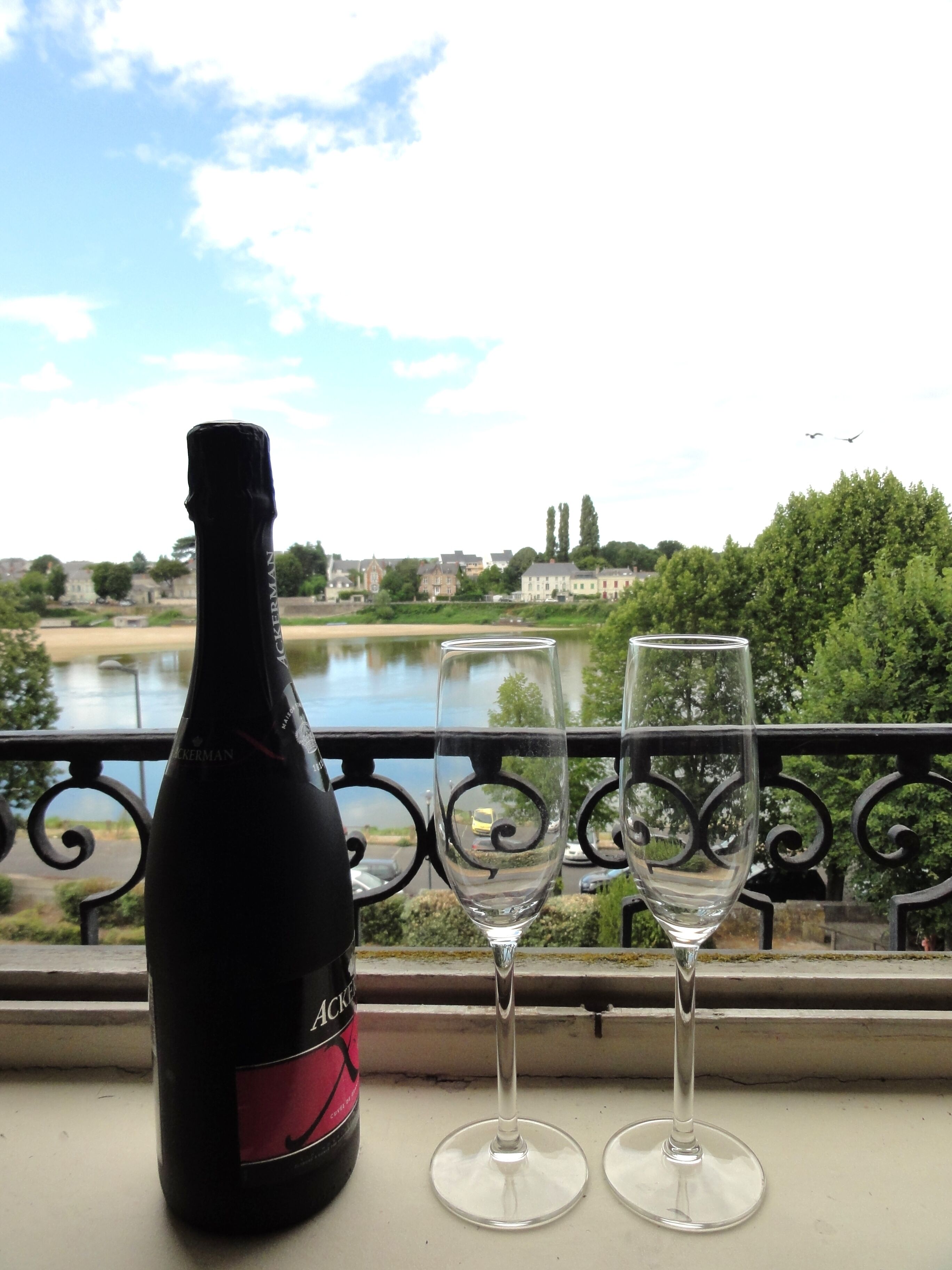 A bottle of wine and two glasses on a balcony with a view of a river and buildings.