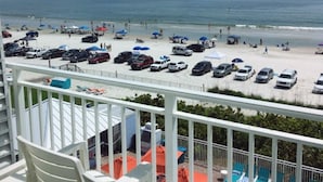 A balcony with white plastic chairs, a pool area with blue lounge chairs, and a view of a beach with parked cars and umbrellas. - Amazing! Direct Oceanfront Condo - Coconut Palms Beach Resort II* Special Rates* (New Smyrna Beach)
