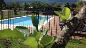 A swimming pool with a wooden deck and a metal fence, surrounded by greenery and a mountainous backdrop.