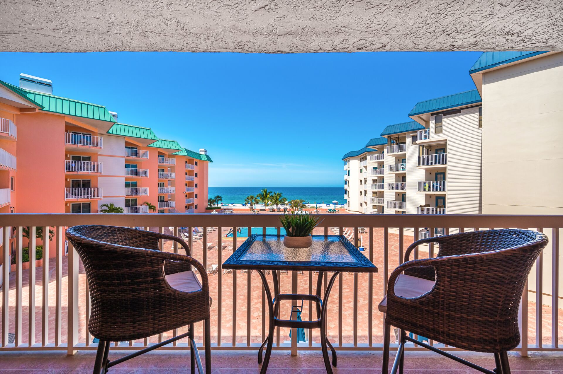 A balcony with wicker chairs and a table, overlooking a beach with buildings in the background.