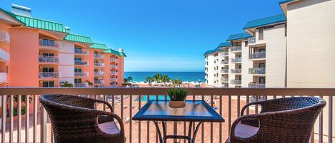 A balcony with wicker chairs and a table, overlooking a beach with buildings in the background.
